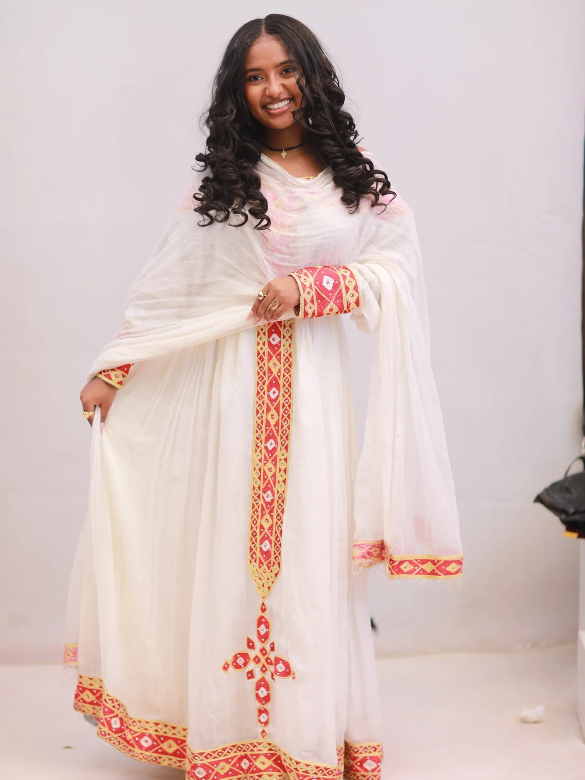 A woman modeling a traditional white Anarkali Habesha kemis with red and gold geometric embroidery, featuring a matching sheer dupatta.
