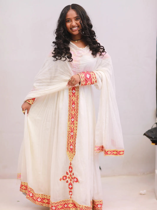 A woman modeling a traditional white Anarkali Habesha kemis with red and gold geometric embroidery, featuring a matching sheer dupatta.