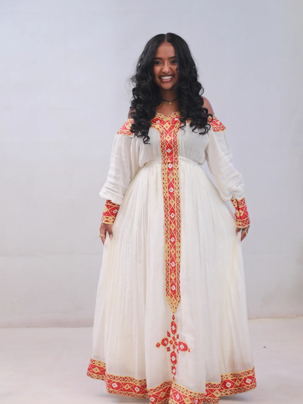 A woman modeling a long off-the-shoulder white Habesha kemis with red and gold embroidered details, standing against a plain light gray studio background.