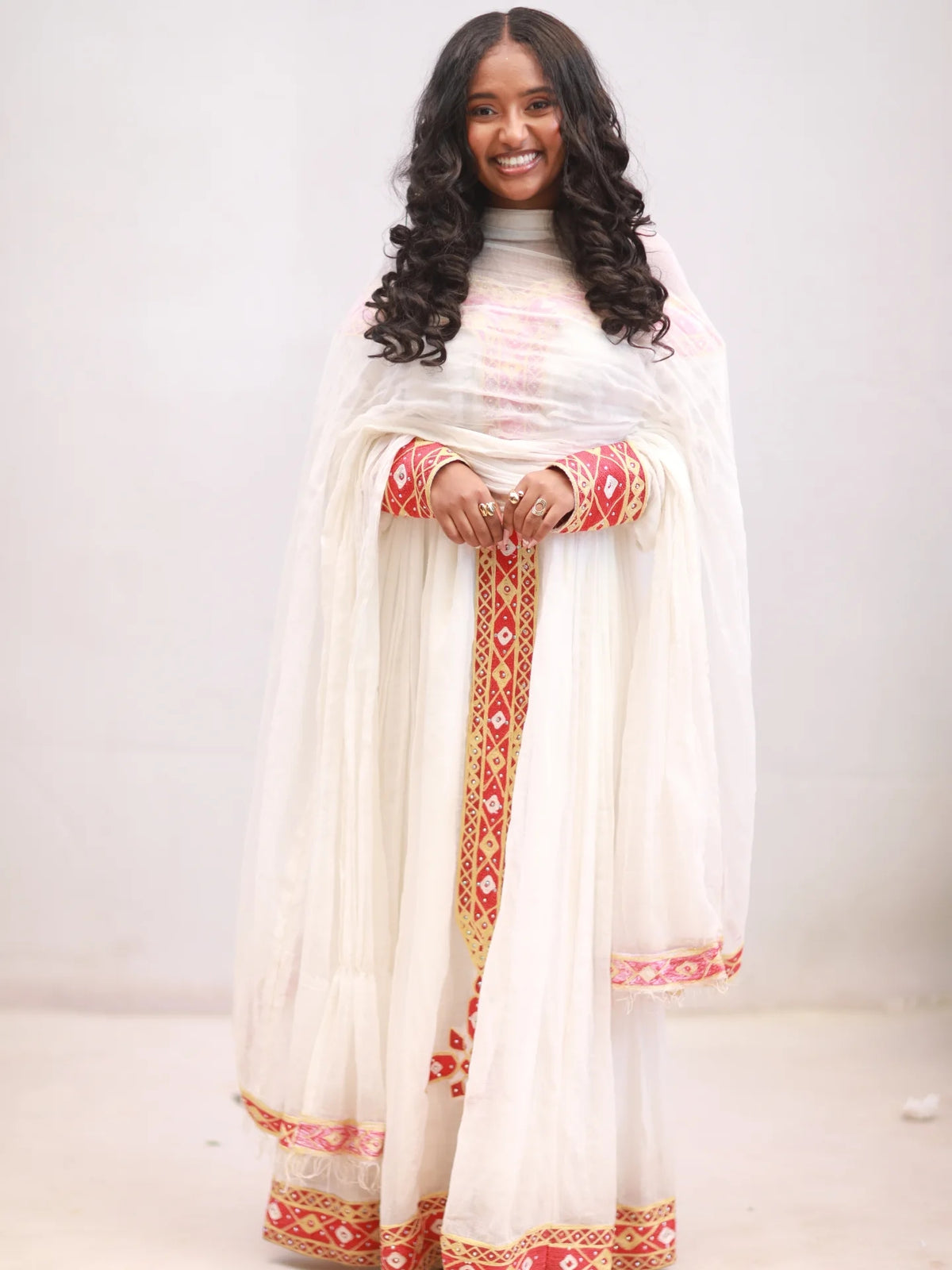 Woman wearing a white traditional Habesha kemis with red and gold patterns on a plain background