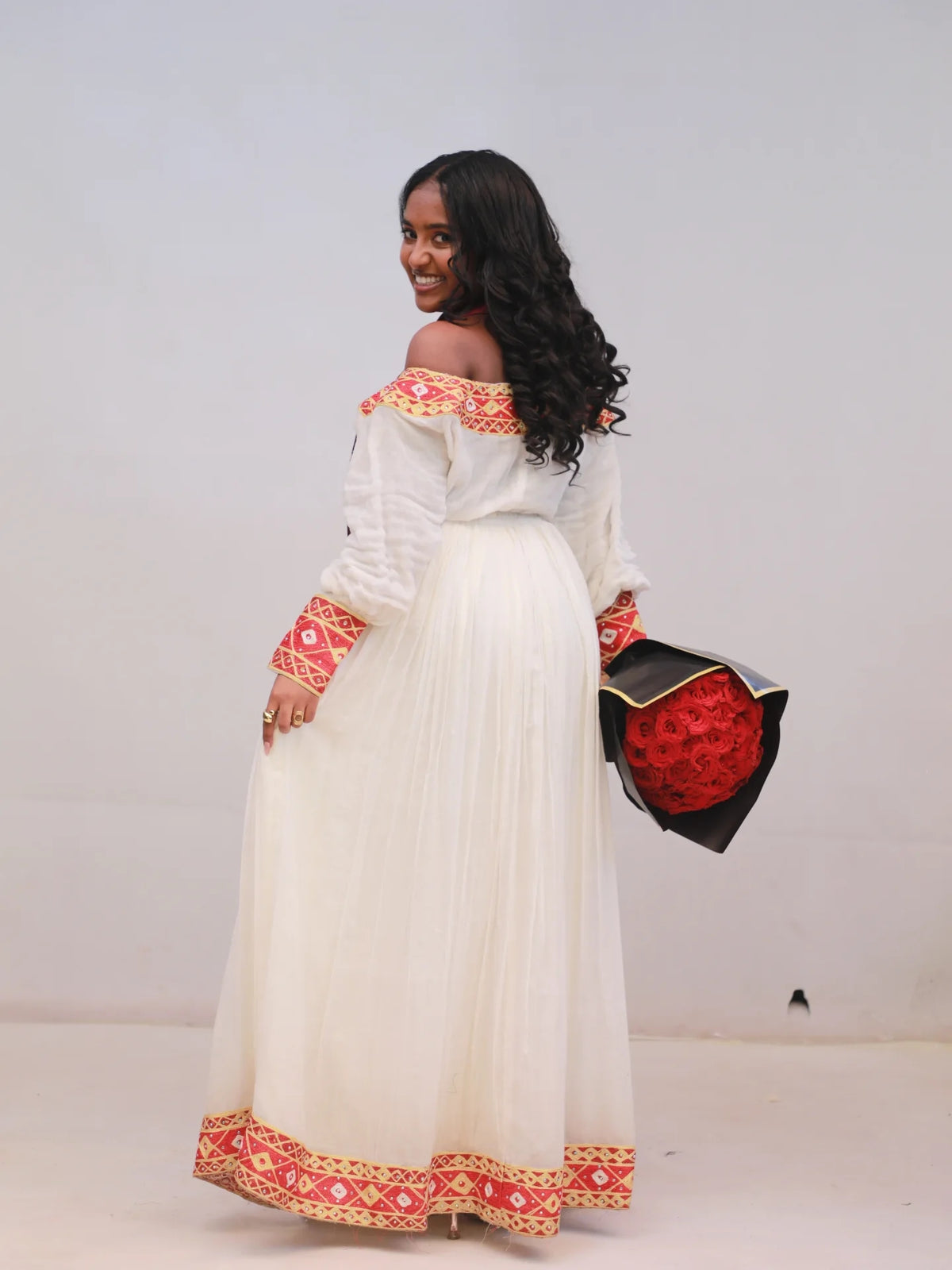 Woman wearing a traditional Habesha kemis with white top and red and orange patterns, holding a black clutch with red floral design.