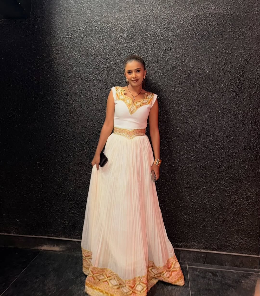 Woman in a white and gold Ethiopian traditional dress standing against a black wall.