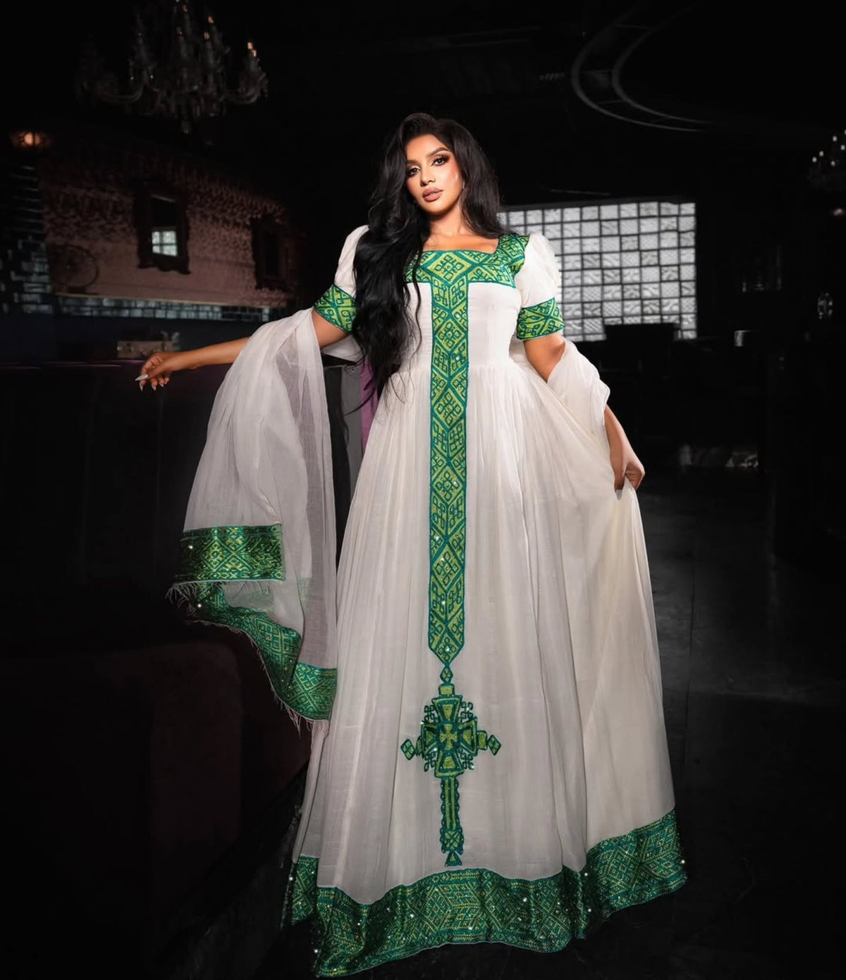 A woman models a traditional Habesha store dress with intricate green embroidery, featuring a long green cross motif on the front and matching green patterned borders on the sleeves and hem, set against a dark, atmospheric background.