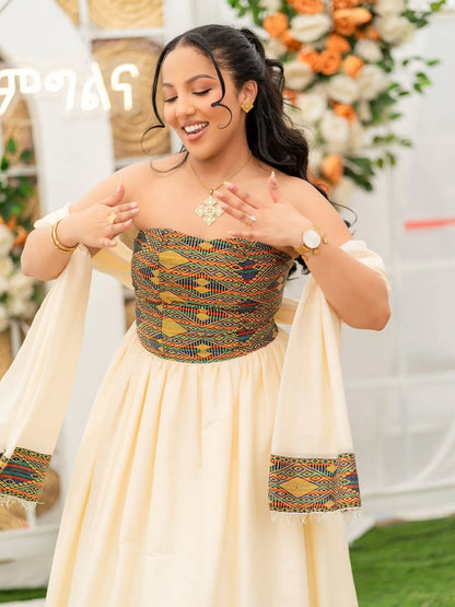A woman wearing a cream-colored traditional Habesha Dress  with a patterned embroidered bodice and matching embroidered cuffs on the sleeves, posing in front of a decorated white arch with floral arrangements.