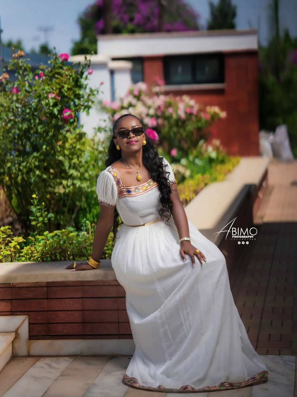 A woman wearing a white Aso Ekwu traditional Ethiopian Dress  with colorful embroidered details on the bodice and hem, posing outdoors in front of a garden and modern building.
