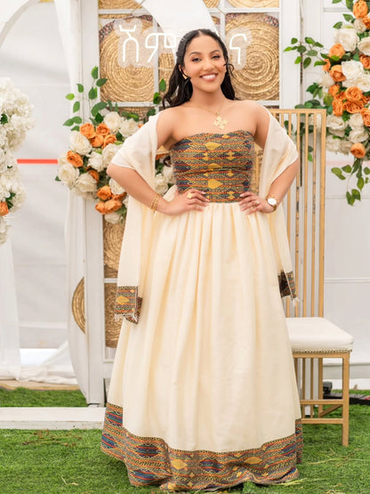 A woman stands outdoors wearing a strapless cream-colored traditional Habesha Dress  with a heavily embroidered gold, brown, and blue patterned bodice and border. She is posing in front of a decorated white arch with floral arrangements and a gold chair.