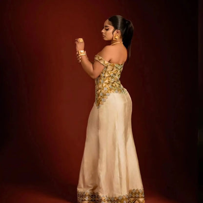 A woman in profile wearing a cream off-the-shoulder ethiopian dress for women with ornate gold embroidery concentrated on the bodice and along the hem, posed against a dark reddish-brown studio backdrop.