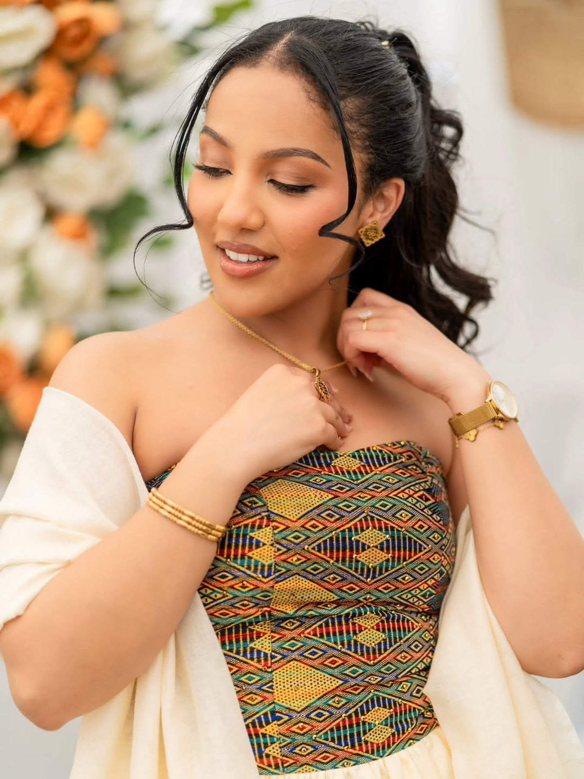 A woman wearing a strapless African wax print Habesha Dress  bodysuit with a colorful geometric pattern in green, yellow, red, and black, styled with a cream-colored shawl and gold jewelry.