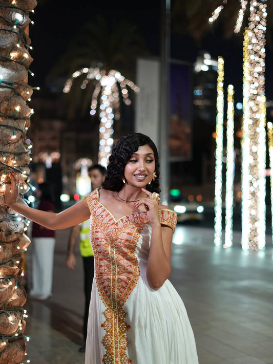 A woman posing outdoors at night wearing a white and orange embroidered traditional Ethiopian Dress for Women , standing next to a decorated palm tree with warm lights.