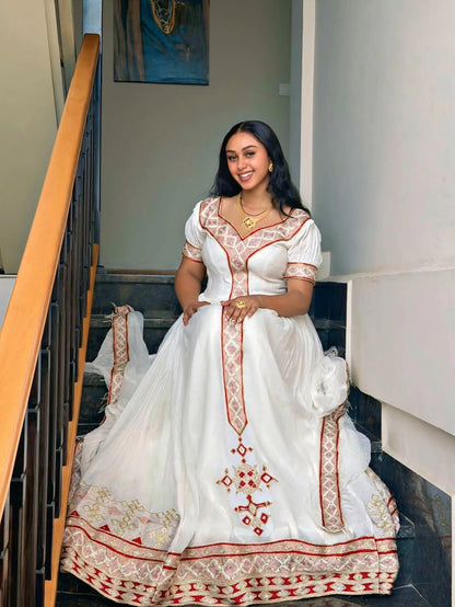 A woman wearing a traditional white embroidered Habesha Kemis  with red and gold detailing, sitting on dark stairs in an indoor setting.