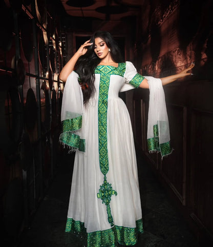A woman modeling a white traditional Habesha store dress with rich green embroidery and patterned borders, posed in a dark, atmospheric corridor.
