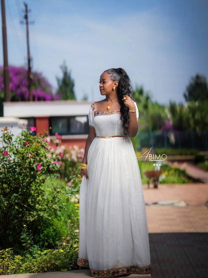 A woman wearing a long white Ethiopian Dress  with embroidered details and a gold waistband, standing outdoors in a garden setting with flowers and greenery in the background.