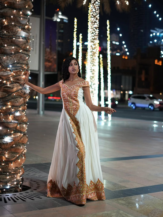 A woman wearing a white off-the-shoulder evening Ethiopian Dress for Women  with gold and orange embroidery, posing outdoors at night next to a decorated palm tree.