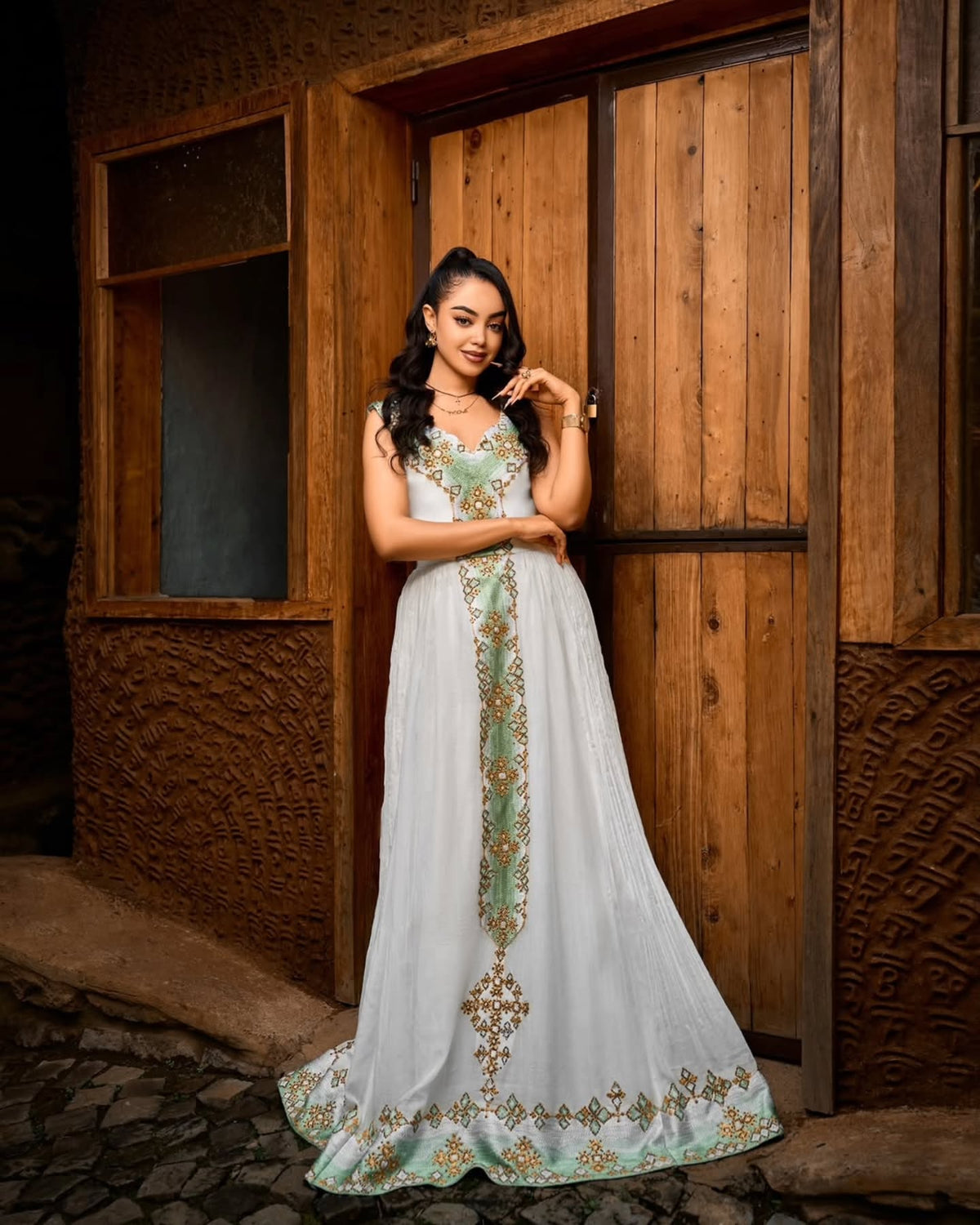 A woman stands in front of a rustic wooden door wearing a long white or off-white formal Ethiopian dress  with mint green and gold embroidery. The dress has a fitted bodice and a flowing skirt with ornate decorative detailing running down the center and along the hem.