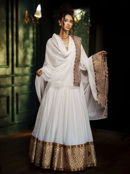 A woman models a traditional white Habesha Kemis with gold patterned borders and a matching draped shawl in a dimly lit interior setting.