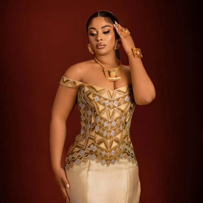 A woman models a gold and beige embellished off-the-shoulder ethiopian dress for women with a cream skirt, accessorized with gold jewelry, against a dark red studio background.