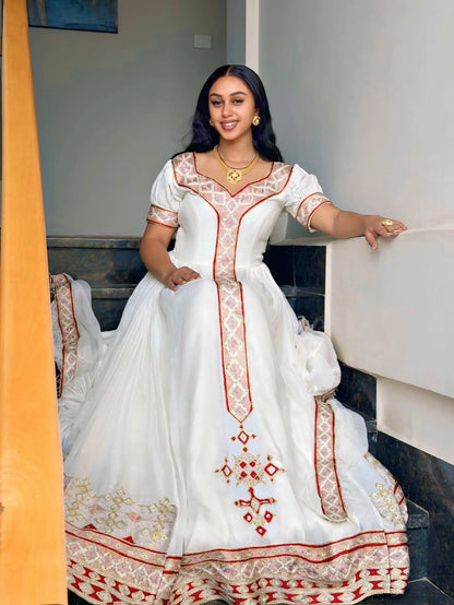 A woman wearing a white traditional Habesha Kemis  with red and gold embroidery, posing on an indoor staircase.