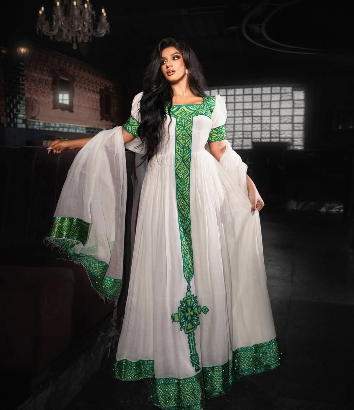A woman modeling a white traditional Habesha store dress with vibrant green embroidered borders and a matching sheer dupatta, set against a dark, elegant interior background.