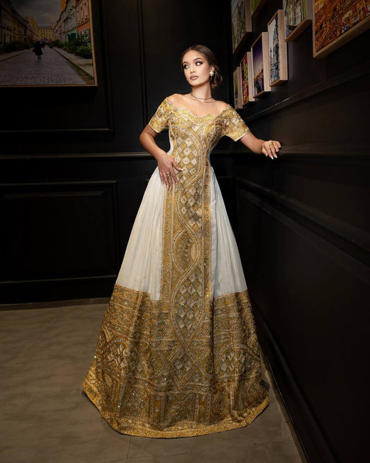 A woman stands in a dimly lit interior wearing an off-the-shoulder formal Woman wearing a traditional embroidered Ethiopia Clothes ses in a dimly lit room with warm lighting. with a cream tulle skirt, heavy red and gold embroidery, and a heavily embellished dark red and gold flared hem.