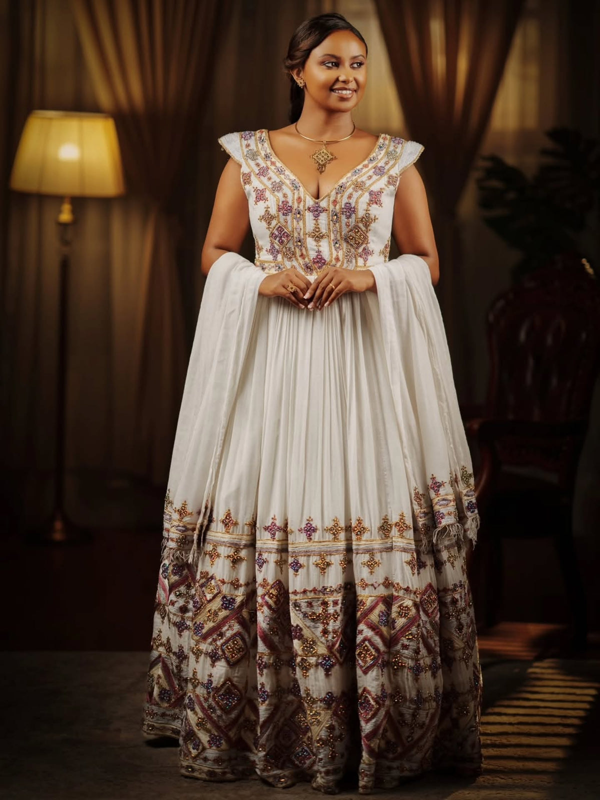A woman wearing a traditional white Ethiopian national dresses with intricate multicolor embroidery and a matching dupatta, standing in a dimly lit room with a lamp in the background.