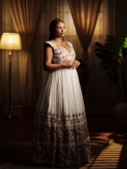 A woman wearing a floor-length white Ethiopian national dresses with floral embroidery and a heavily embellished border stands in a dimly lit room with curtains, a lamp, and a plant.