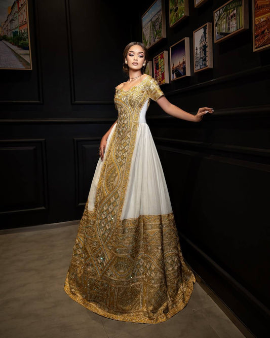 A woman stands in a dimly lit interior wearing an off-the-shoulder formal Woman wearing a traditional embroidered Ethiopia Clothes ses in a dimly lit room with warm lighting. with a cream tulle skirt, heavy red and gold embroidery, and a heavily embellished dark red and gold flared hem.