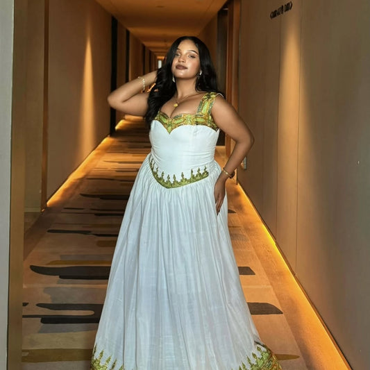 A woman wearing a white sleeveless maxi Habesha kemis with gold embroidery, standing in a hotel hallway.