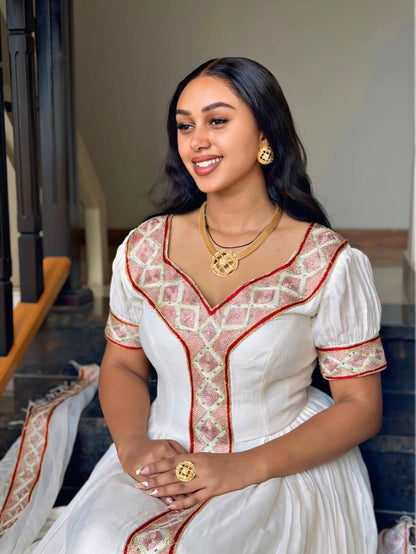 A woman wearing a white traditional Habesha Kemis  with pink and gold embroidered patterns, seated on dark steps. She is accessorized with gold jewelry, including a necklace, earrings, and a ring.