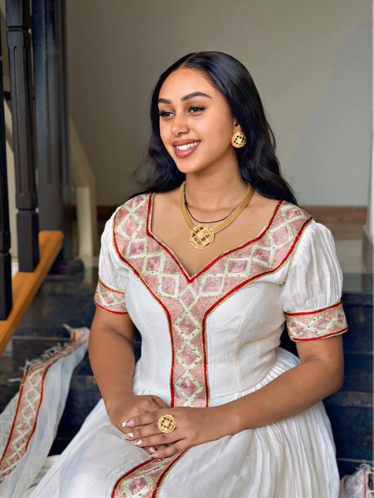 A woman wearing a white traditional Habesha Kemis  with pink and gold embroidered patterns, seated on dark steps. She is accessorized with gold jewelry, including a necklace, earrings, and a ring.