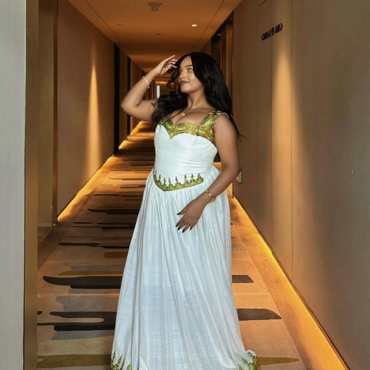 A woman wearing a white sleeveless maxi Habesha kemis with gold embroidery stands in a hotel hallway.