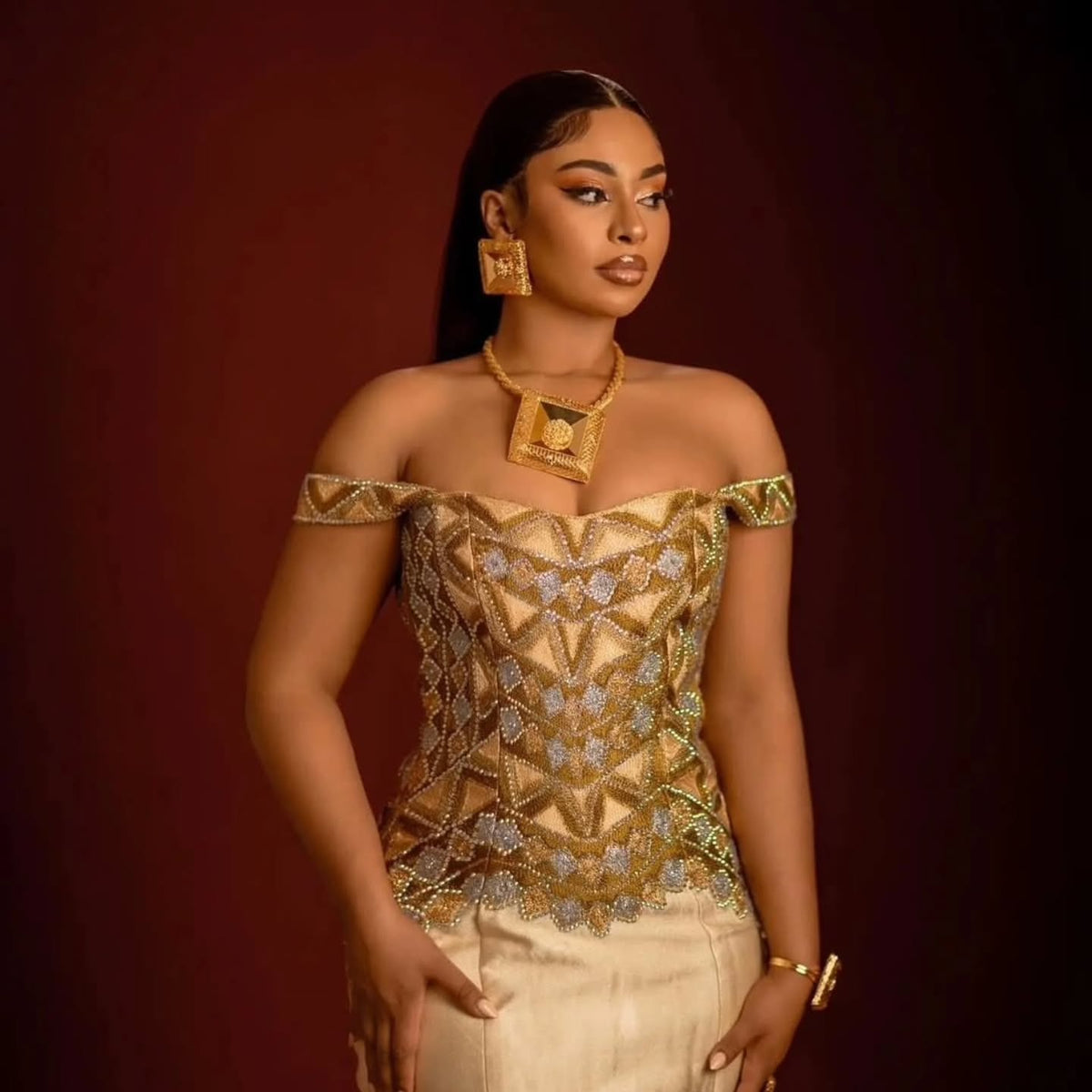 A woman models an off-the-shoulder gold and silver beaded ethiopian dress for women with a geometric pattern, paired with cream-colored pants, set against a dark burgundy background.