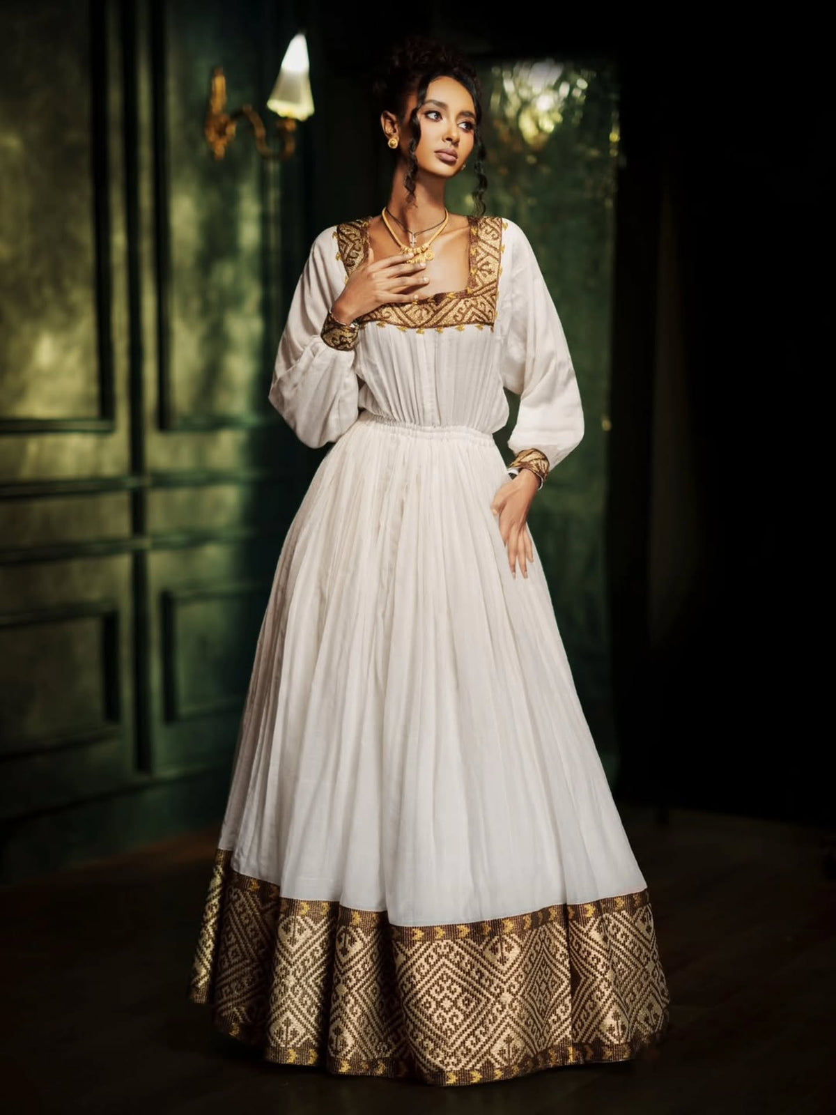 A woman models a traditional white Habesha Kemis dress with ornate gold embroidery on the neckline, sleeves, and hem, set against a dark green paneled background.