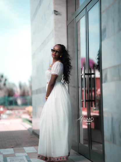 A woman wearing a white puff-sleeve Ethiopian Dress  with a patterned hem and sunglasses, posing outdoors next to a glass door on a stone building.