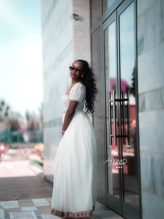 A woman wearing a white puff-sleeve Ethiopian Dress  with a patterned hem and sunglasses, posing outdoors next to a glass door on a stone building.