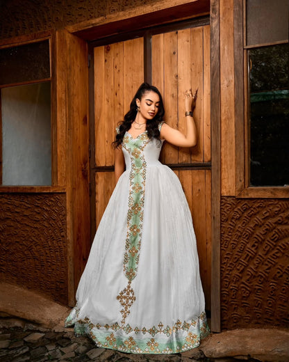 A woman wearing a white traditional Ethiopian dress  with green and gold embroidery stands in front of rustic wooden doors.