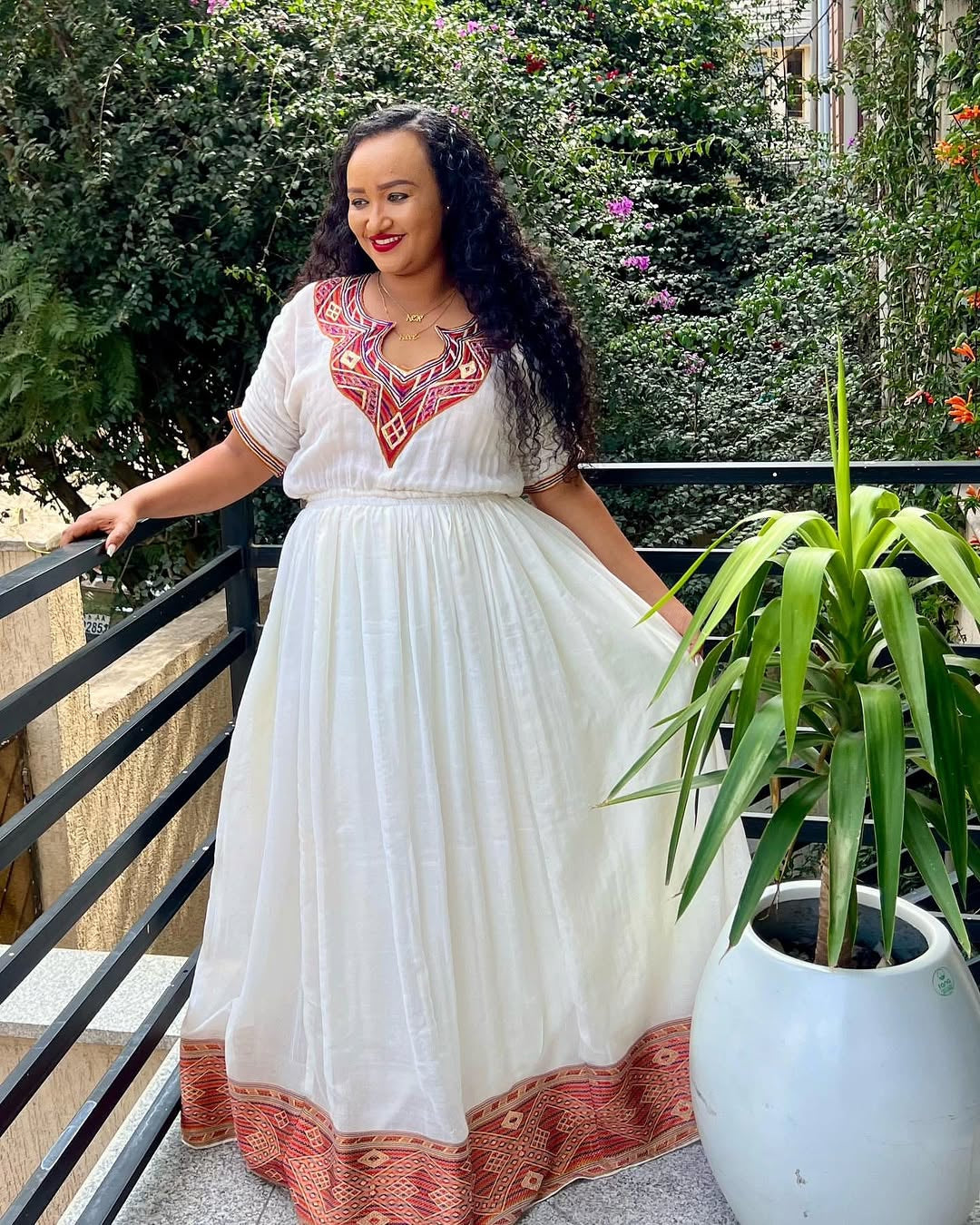 Woman in a white dress with Ethiopian Dress embroidery standing on a balcony with plants.