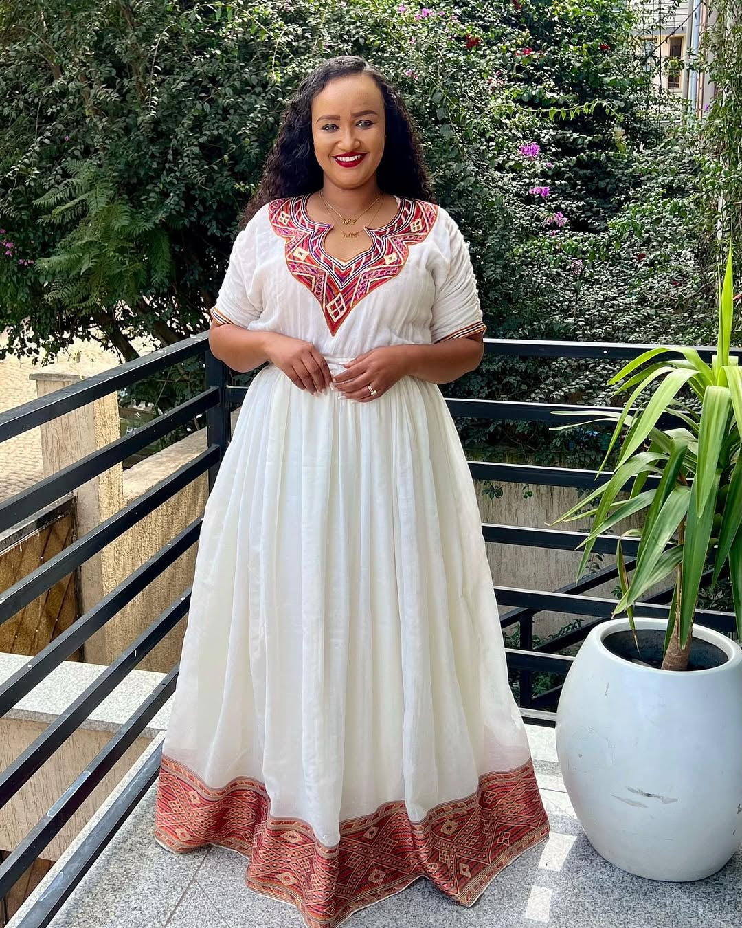 Woman in a white Ethiopian Dress with red embroidery standing on a balcony with greenery.