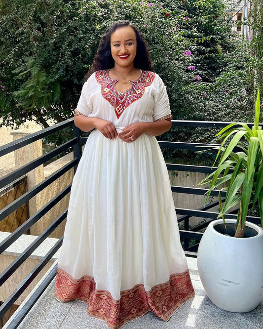 Woman in a white Ethiopian Dress with red embroidery standing on a balcony with greenery.