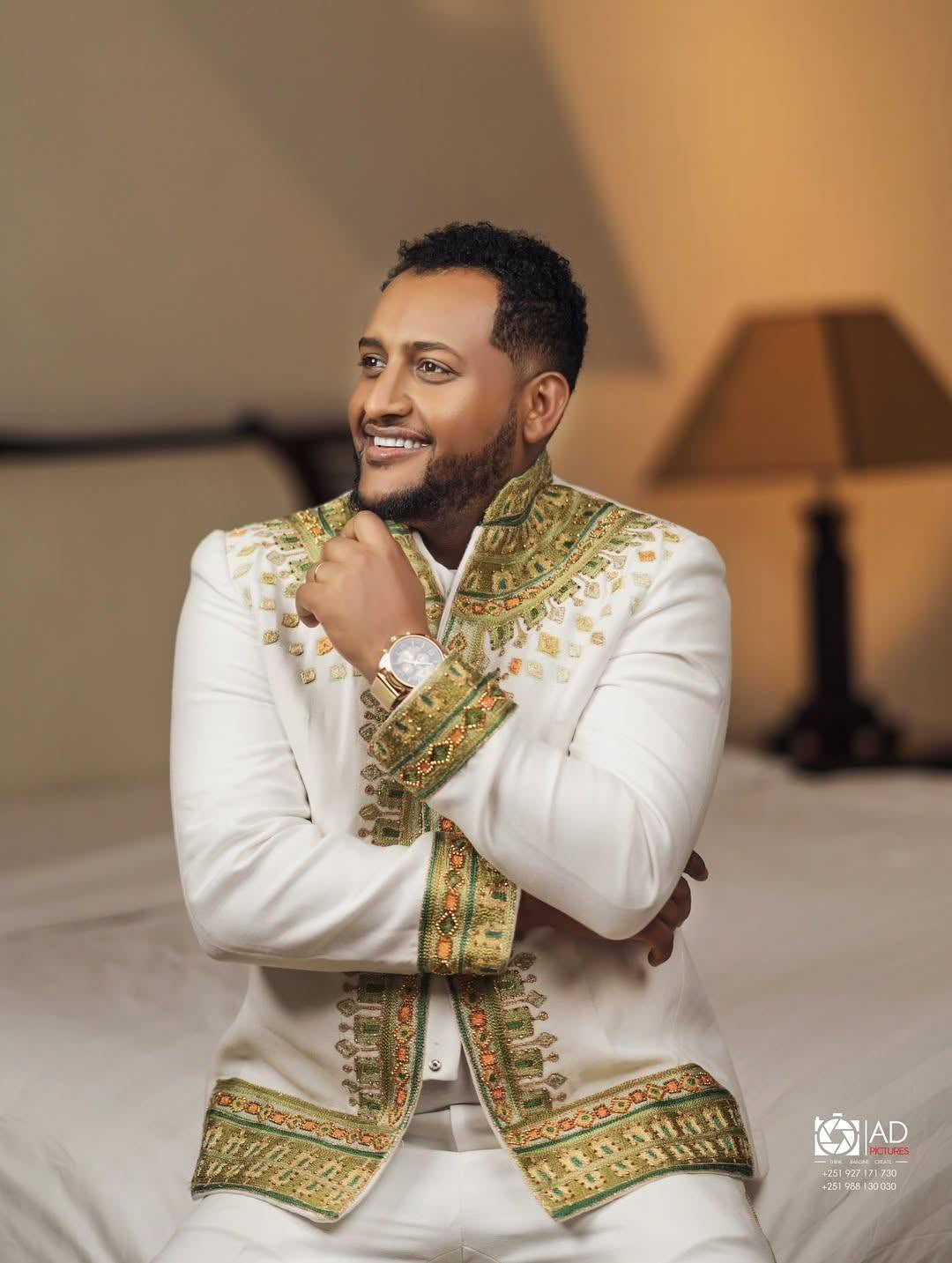 Man wearing a white and green Habesha Men’s Wedding Outfit embroidered jacket sitting indoors.