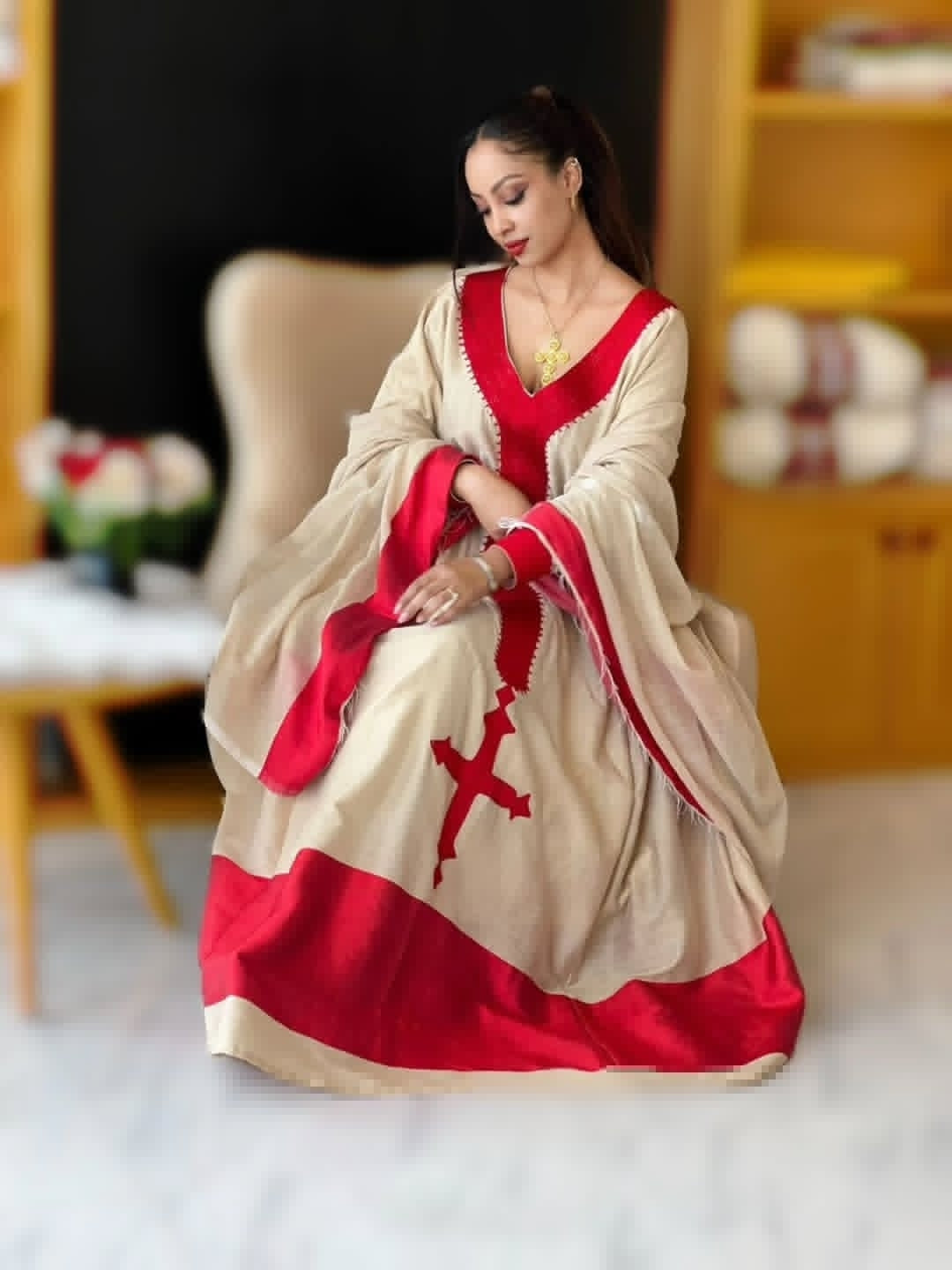 Woman in a red and beige Ethiopian traditional dress with a cross design sitting on a chair.