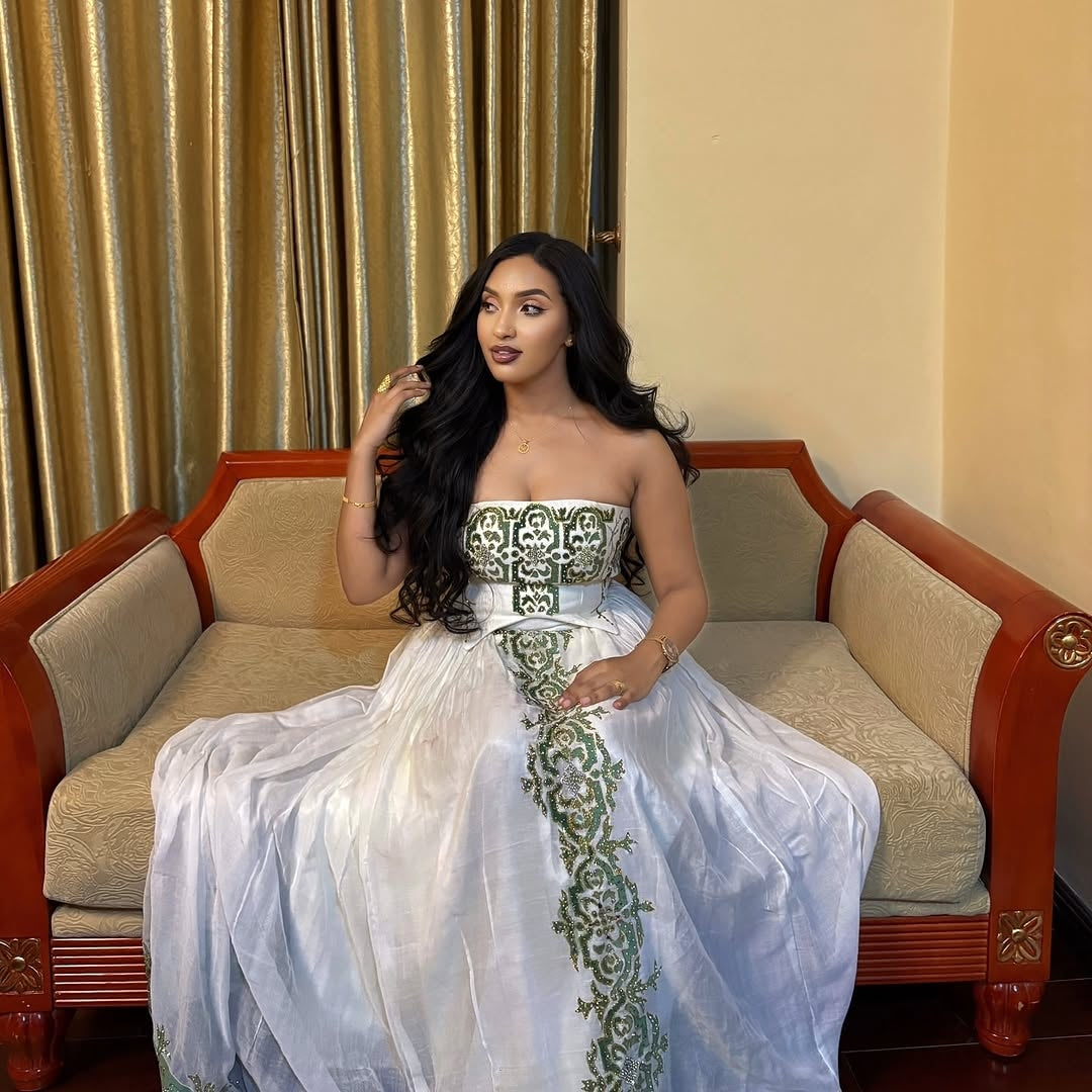 Woman in a white and green Ethiopian dress sitting on an ornate couch with gold curtains in the background