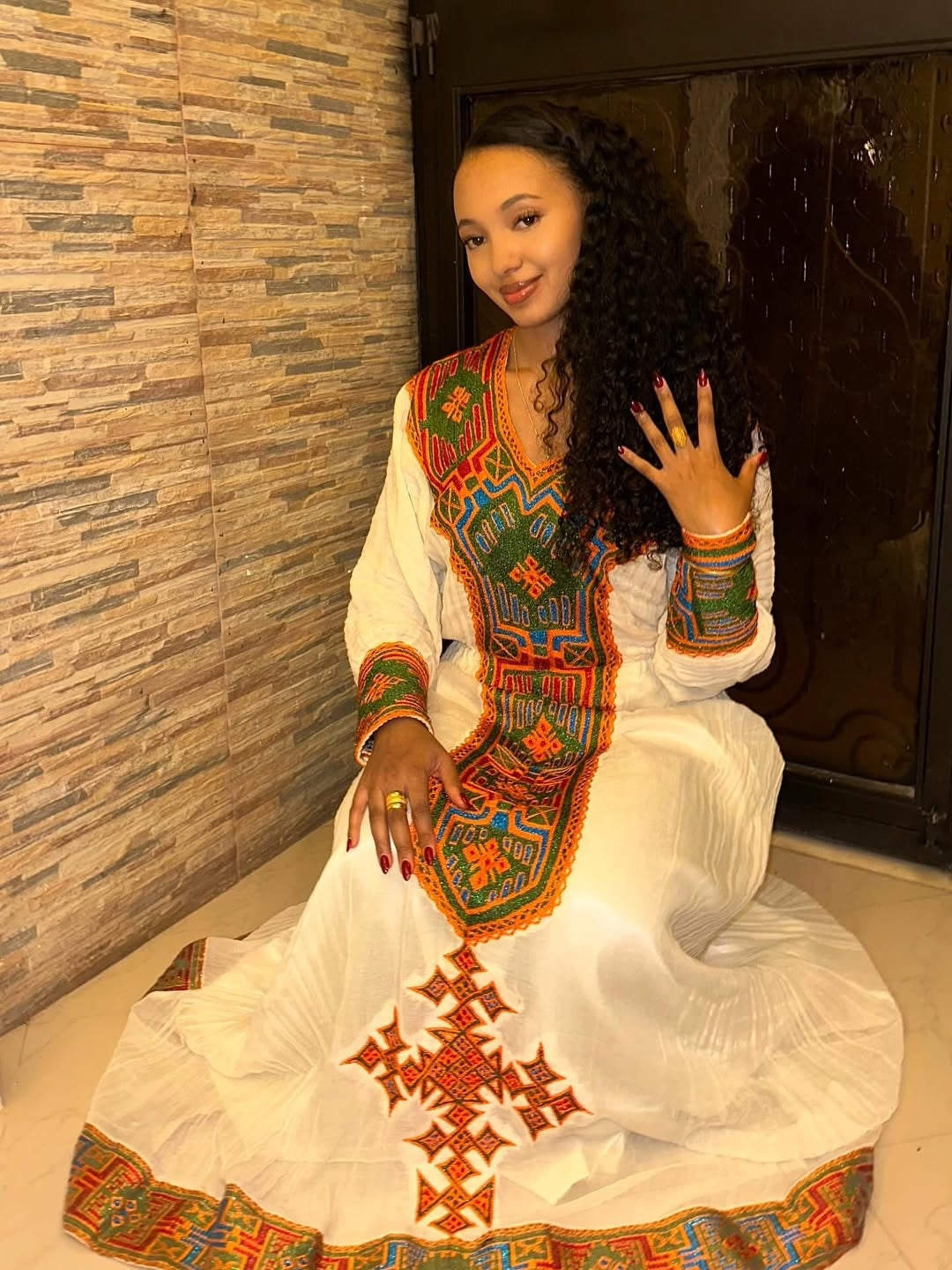 Woman in a Habesha Libs with colorful patterns sitting against a stone wall.
