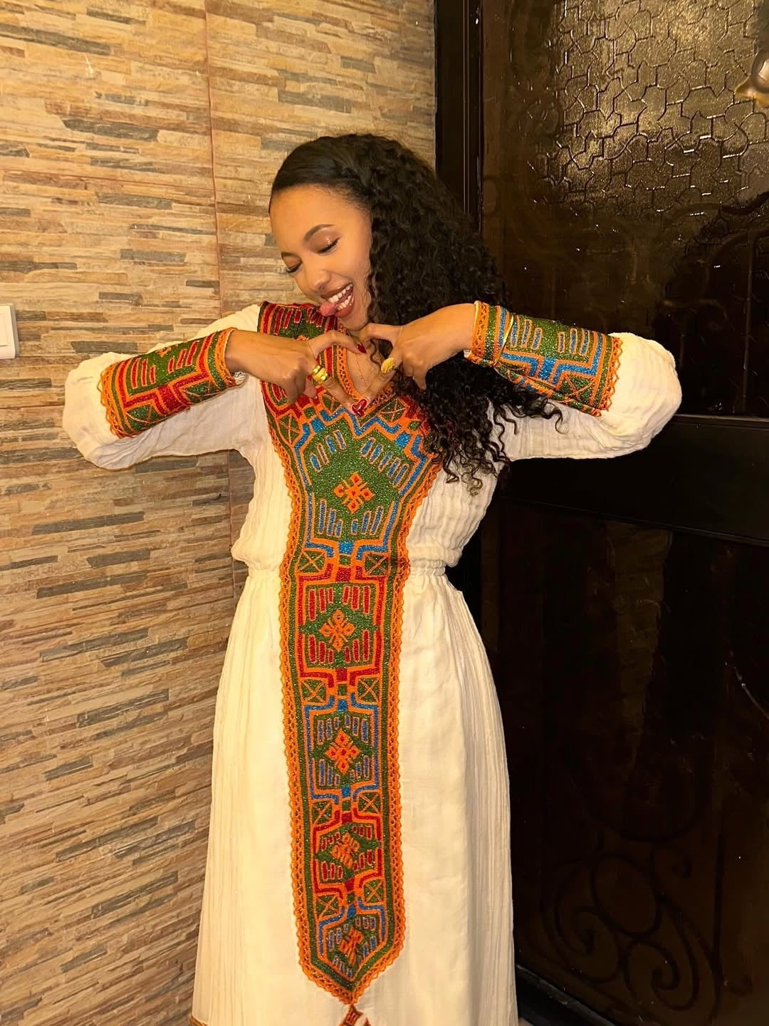 Woman wearing a Habesha Libs embroidered dress with a colorful pattern against a textured wall.