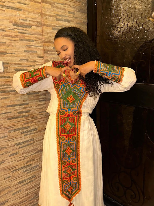 Woman wearing a Habesha Libs embroidered dress with a colorful pattern against a textured wall.