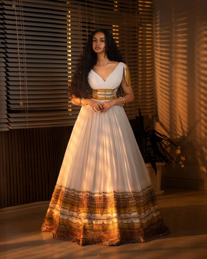 Woman in a white and gold traditional outfit standing in a softly lit room.