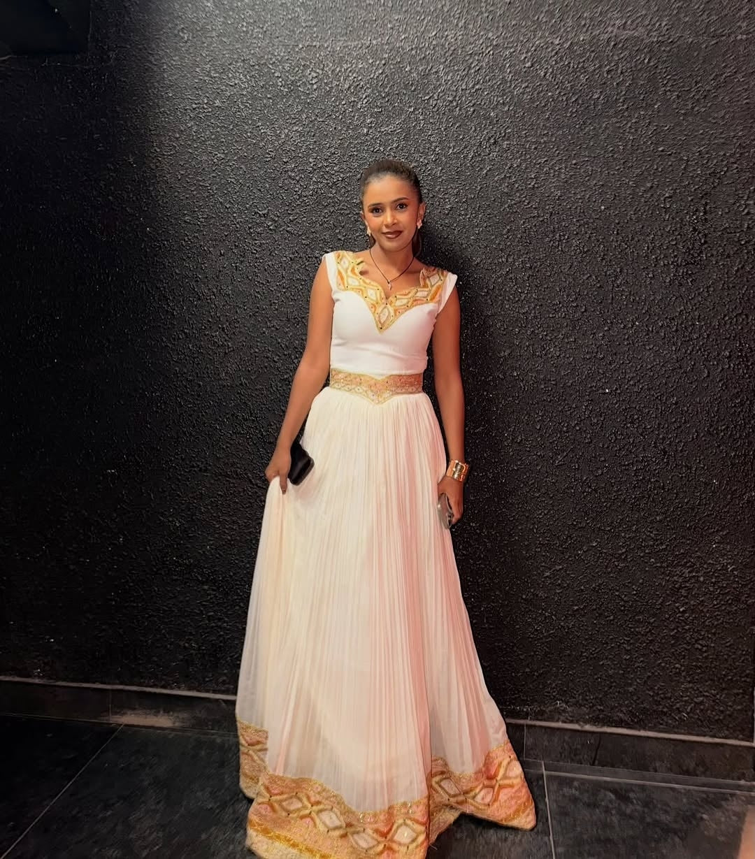 Woman in a white and gold Gold Ethiopian Kemis standing against a black wall.