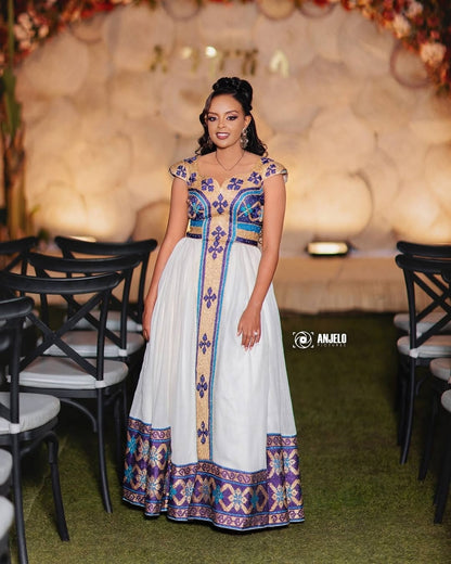 Woman in a colorful Ethiopian Traditional Dress standing in an elegant indoor setting with decorative chairs and floral arrangements.
