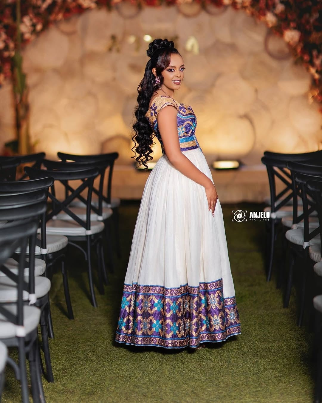 Woman in a colorful dress standing in an elegant indoor setting with floral decorations.