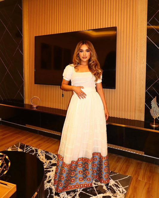 Woman in a white Ethiopian Dress  with colorful patterns standing in a modern room.