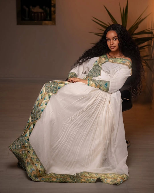 Woman in a white and green traditional Ethiopian libs  sitting indoors with a plant in the background
